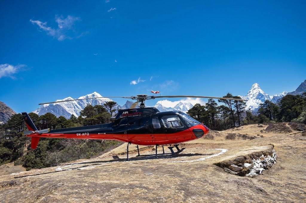 Red and black helicopter parked on a mountain helipad with the snow-capped peak of Mount Ama Dablam and Himalayan pine forests in the background.